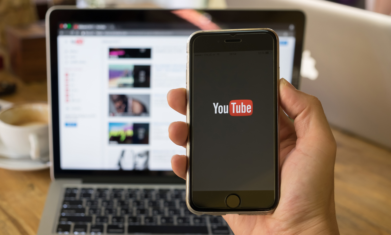 someone holding a mobile phone loading youtube with a laptop on youtube in the background